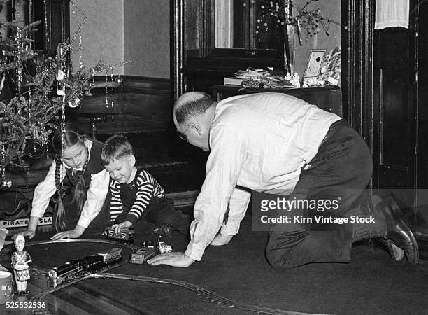 On his hand and knees, a father plays with his daughter and son with a model train under the Christmas tree.