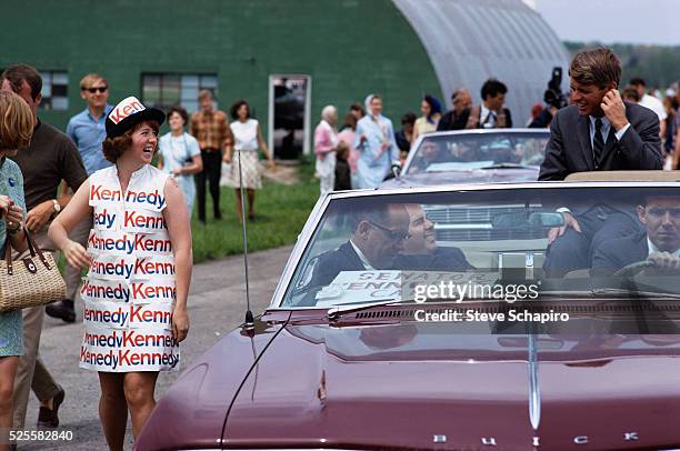 Robert Kennedy smiles at a supporter who walks beside his car in a dress made of Kennedy campaign posters.