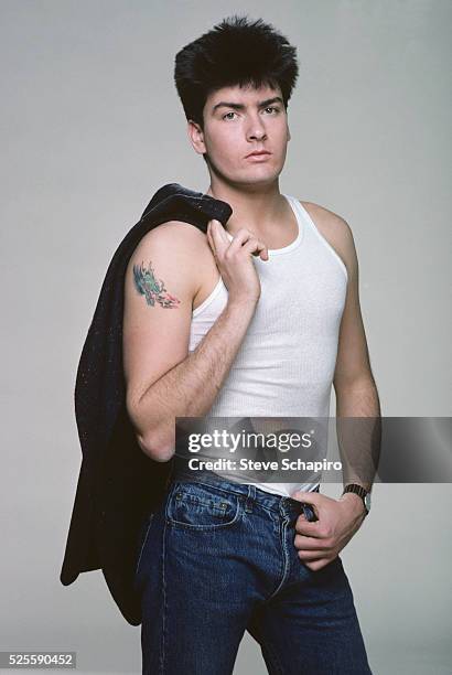 American actor Charlie Sheen wearing a white vest and blue jeans, his jacket slung over his shoulder, in a studio portrait, circa 1985.