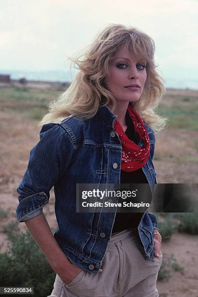 American actress and singer Beverly D'Angelo wearing a blue denim jacket over a black top and a red neck scarf, her hands in the pockets of her beige...