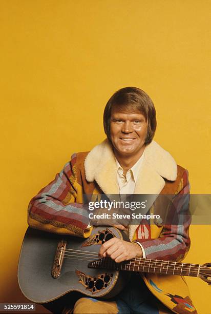 American singer-songwriter and guitarist Glen Campbell poses wearing a patterned fleece-lined coat, holding his guitar in a studio portrait, against...