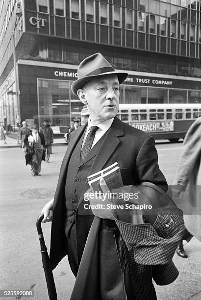 British actor Alec Guinness wearing a three-piece suit, a fedora and a tie, holding an umbrella and carrying a coat and a books in his arm, as he...