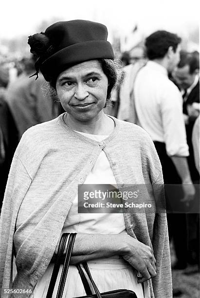 American civil rights activist Rosa Parks wearing a dark hat, a cardigan over her shoulders, with her arm looped through the straps of a handbag,...