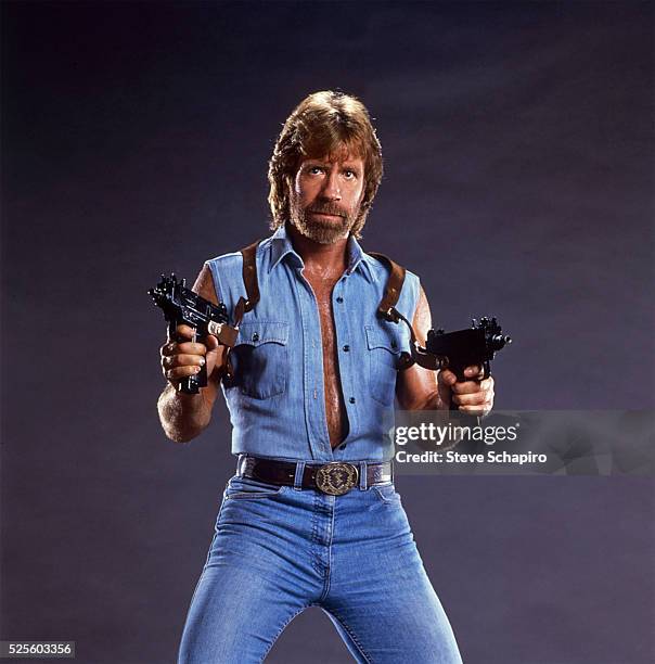 Chuck Norris poses with two Uzis, his sleeveless denim shirt unbuttoned to his waist. Publicity for Invasion USA