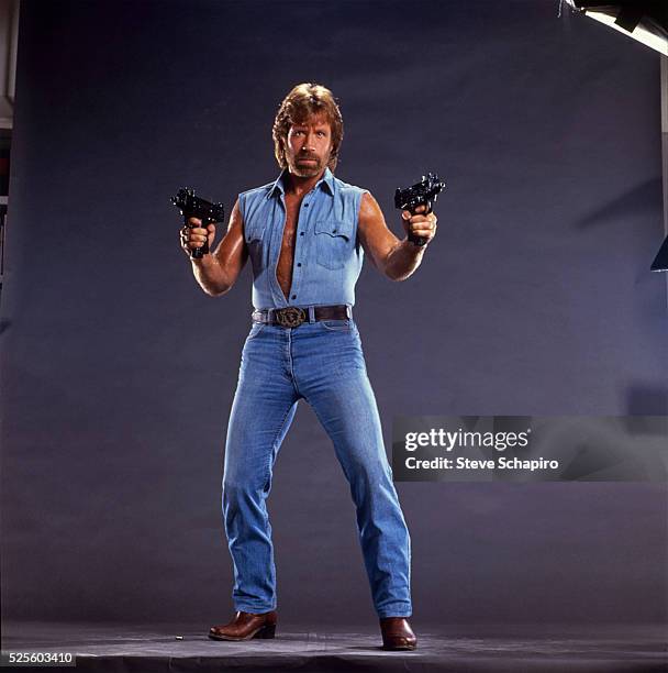 Chuck Norris poses with two Uzis, his sleeveless denim shirt unbuttoned to his waist.Publicity for Invasion USA.