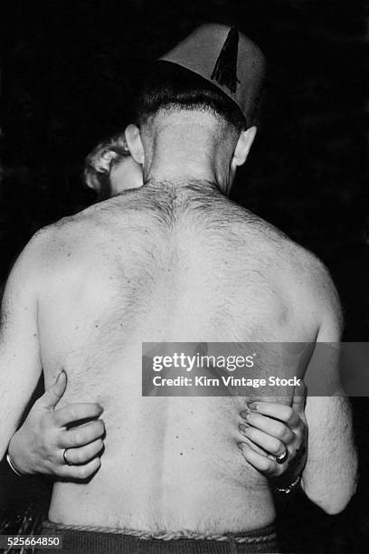 Man with a hairy back and wearing a fez kisses a woman with her long finger nails impaling his skin.