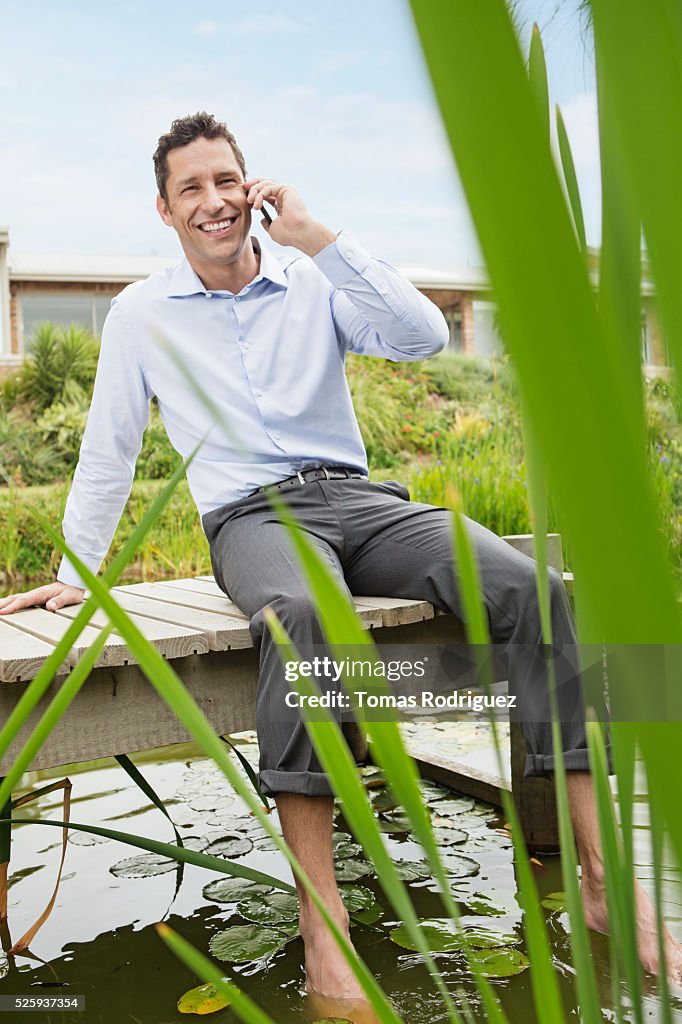 Man sitting on jetty by pond and talking on cell phone