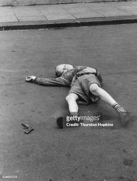 Little boy with a toy gun lies in the road after being 'shot' by a friend, 7th August 1954. Original Publication : Picture Post - 7230 - Children Of...