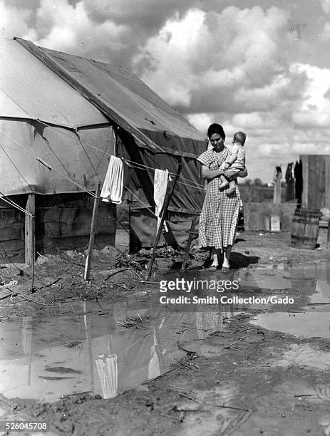 Black and white photograph of a woman holding an infant, walking in a wet, muddy camp, "titled "Migrants' Camp", by Dorothea Lange, American...