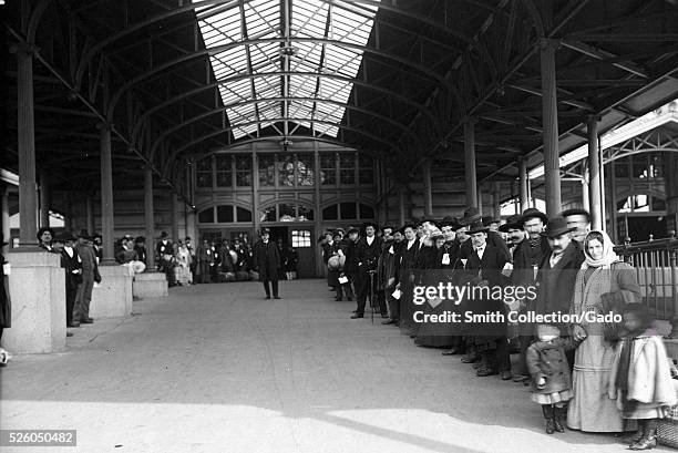 Black and white photograph of a large group of immigrants lined up outside waiting to be taken off Ellis Island, by Edwin Levick, Ellis Island, New...