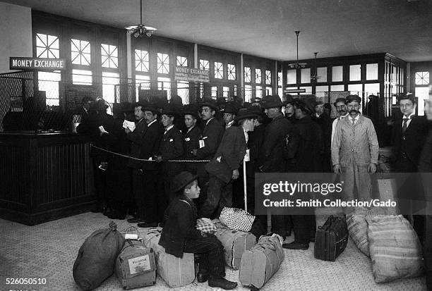 Black and white photograph of a large group of immigrants with baggage lined up at tellers windows marked "Mone Exchange", by Edwin Levick, Ellis...