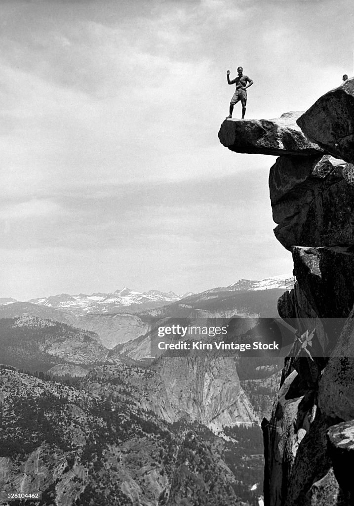 A man waves while perched dangerously on an overhanging rock. Yosemite, ca. 1920