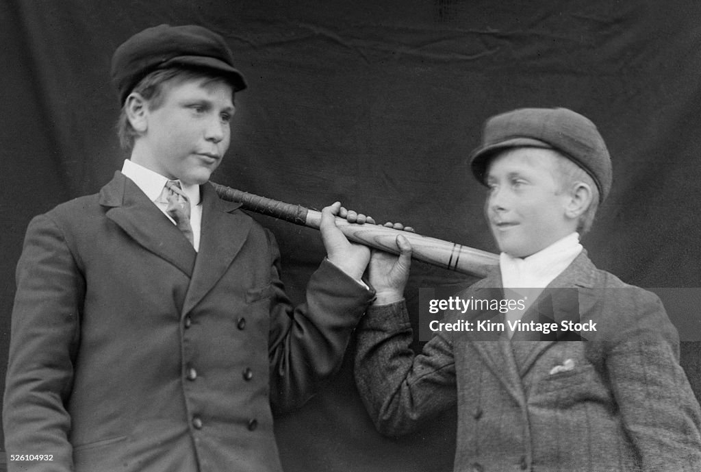 Two young boys pose with a baseball bat in a studio shot, ca. 1905