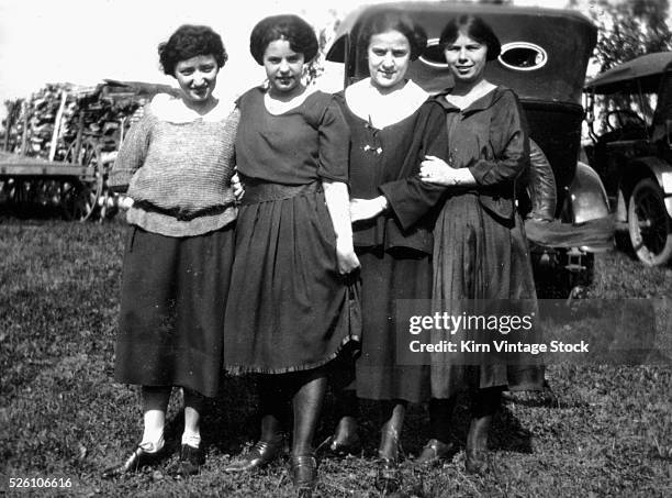 Four young women stand in front of parked cars, ca. 1920