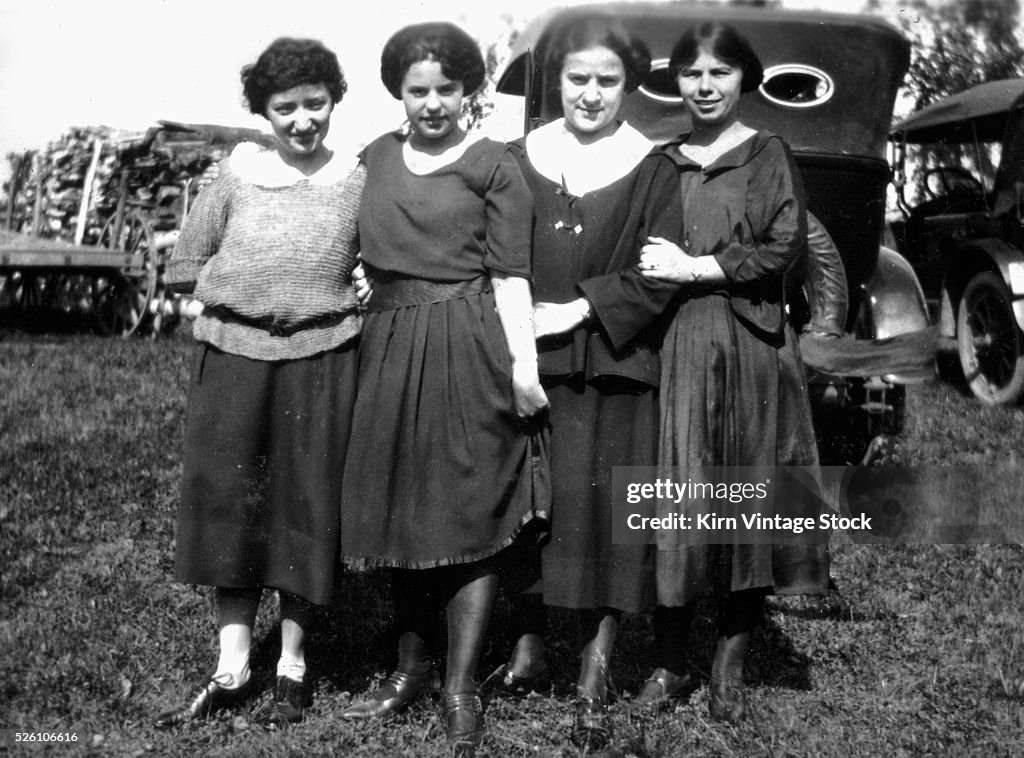 Four young women stand in front of parked cars, ca. 1920