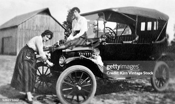 Young woman cranks the car while a girlfriend and a little boy wait.