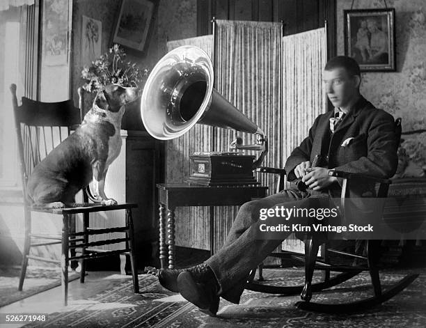 Man holding a harmonica and beagle sitting on a chair listen to the sounds coming from a cylinder phonograph. The photograph recalls the trademarked...