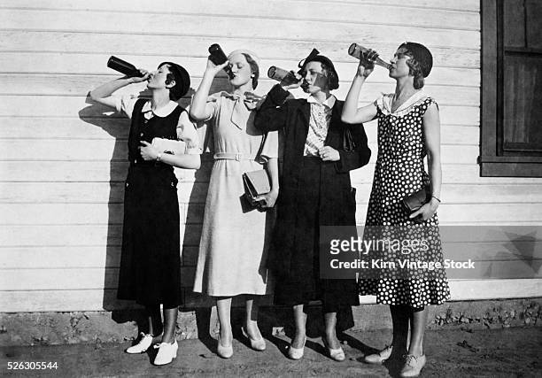 Four women line up along a wall and chug bottles of liquor in the 1920s.
