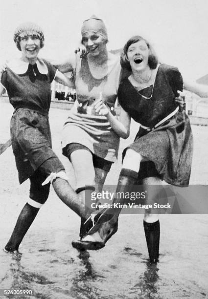 Three women in period swimwear kick up their legs together and laugh while wading in the ocean.