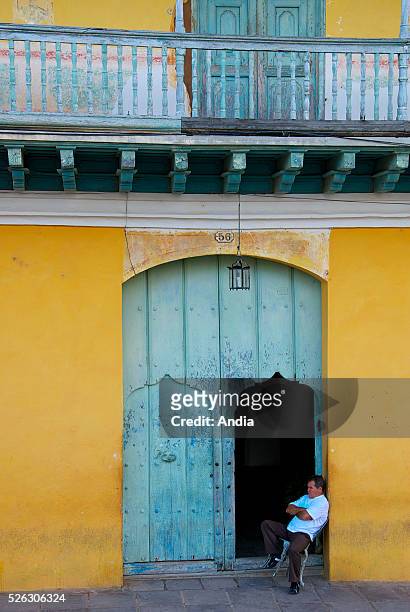 Casa Ortiz or Galeria de Arte , Plaza Mayor, man seated in front of the Colonila mansion, Trinidad, Cuba. . Yellow and blue.