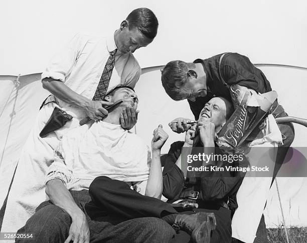 Using a kitchen knife and a pair of pliers two young playfully pretend to remover facial hair from the faces of two other guys in front of their tent...