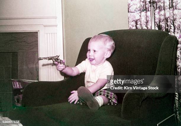 Young boy wielding a toy gun plays in the family easy chair next to the fireplace.