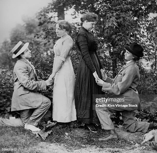 Two women on bended knee and dressed as men, appear to be proposing marriage to two women in unison, circa 1910.