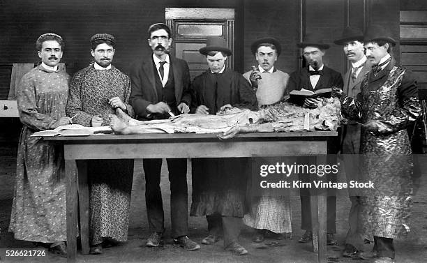 Group of doctors in training in the late 19th century examine a dissected human body laid out on a wooden table.