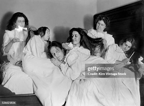 Group of six young women lounge in one bed and work on each others hair.