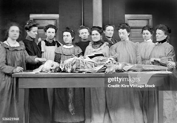Group of women in nurses training examine the dissected remains of man laid out on a table.