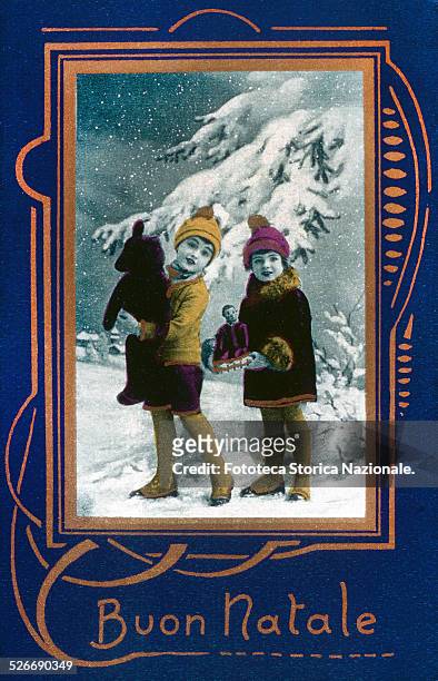 Two children in a snow-covered landscape, under a spruce load of snow, holding a teddy bear and a mechanical doll, just received as a gift. An...