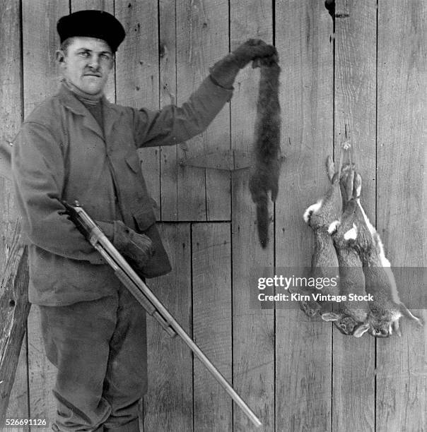 An armed hunter holds up a mink pelt while three dead rabbits hang around in the background.