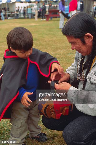 Quinault man helps his young son, named James Black and Tow Wah Us, with a cedar ceremonial mask. The boy also wears a traditional blanket. Quinault...
