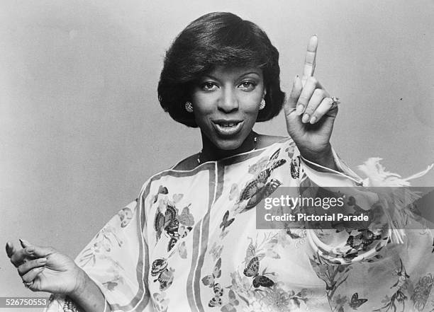 Portrait of singer Natalie Cole, pointing her finger and smiling, wearing a dress with a flower and butterfly pattern, circa 1975.