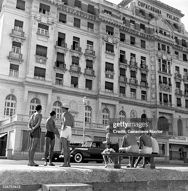 Young men in swimsuits sit in the sun outside the Copacabana Palace, a posh hotel at Copacabana Beach.