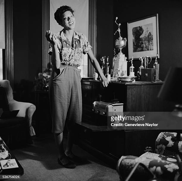 Tennis champion Althea Gibson stands beside the Wimbeldon trophy in her living room.