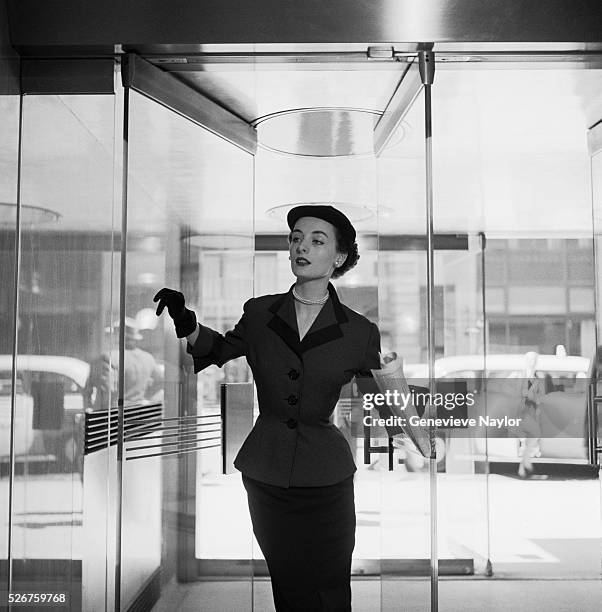 Fashion model wears a wool day suit by Hattie Carnegie, as she walks through a glass doorway.