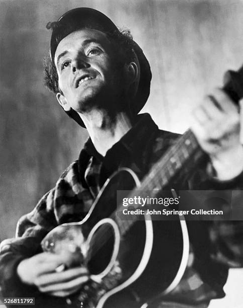 Waist-up photo of American folk singer and composer Woody Guthrie playing his guitar. Ca. 1960s.