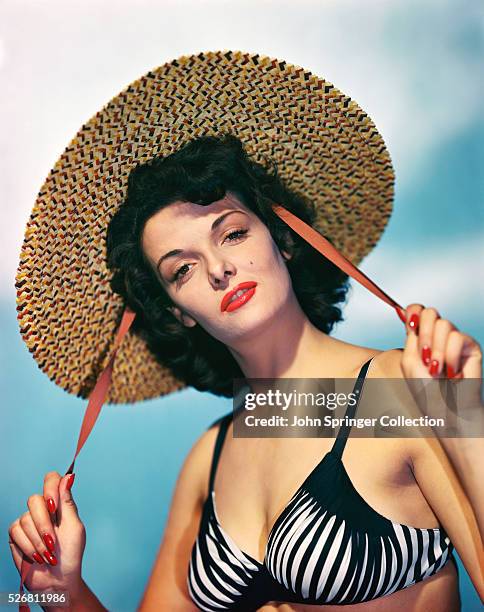Actress Jane Russell wearing a black and white bikini top while holding the red draw strings of a wide brim straw hat. Color publicity handout,...