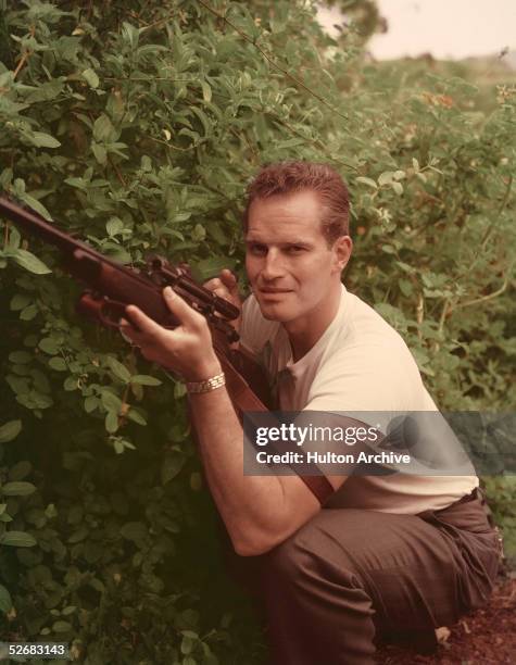 American actor Charlton Heston crouches next to a bush with his rifle raised, butting against his chest, early 1960s. Heston would be elected...