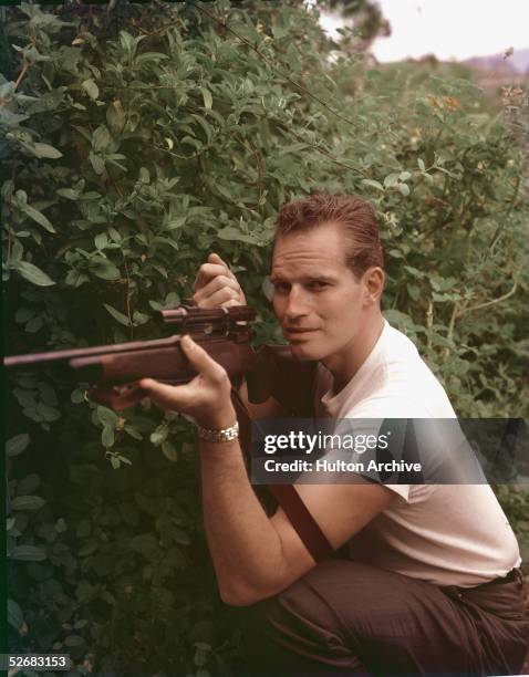 American actor Charlton Heston crouches next to a bush, cocking the trigger of his raised rifle, early 1960s. Heston would be elected President of...