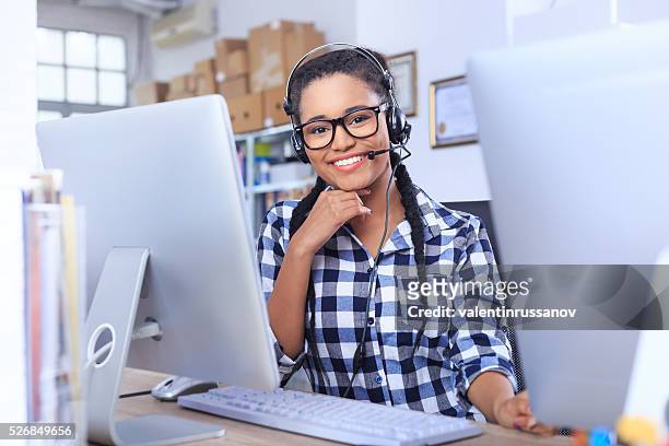 mulher jovem sorridente com auscultadores com microfone sentado no local de trabalho - call center agents imagens e fotografias de stock