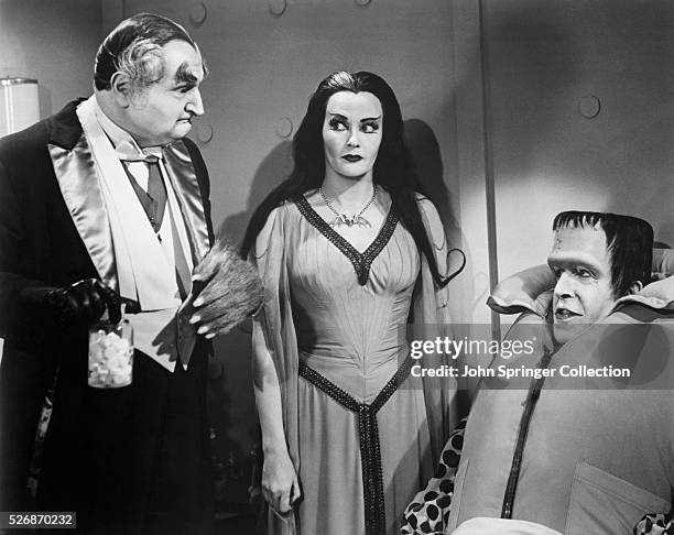 Grandpa holding pills, Lily eyeing Grandpa, and Herman wearing a life vest during an episode of The Munsters.