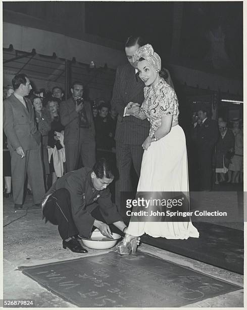Man kneels to help Carmen Miranda make a footprint in the cement at Mann's Chinese Theater in Hollywood, California.