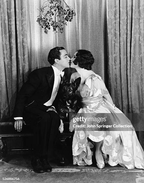 Actor Buddy Rogers and actress Mary Brian kiss under the mistletoe with Baron the German shepherd between them.