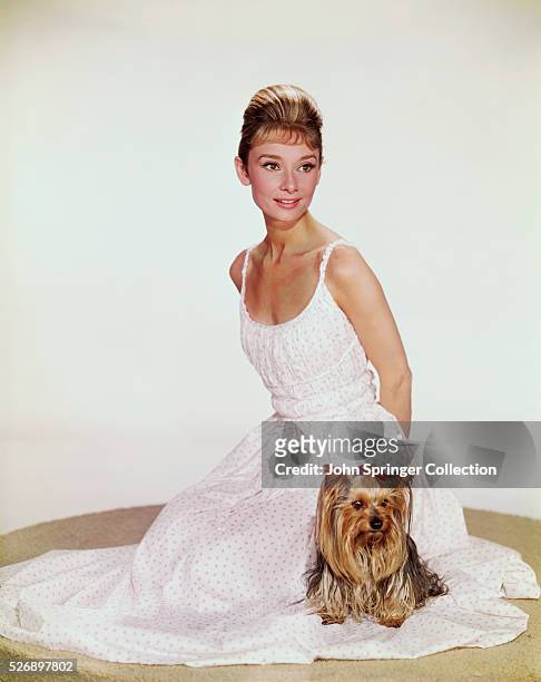 ACTRESS AUDREY HEPBURN IN A DEMURE SLEEVELESS SUMMER DRESS. SHE IS SITTING ON A PLATFORM; THERE IS A CUTE PUPPY SITTING ON THE SKIRT OF HER DRESS....