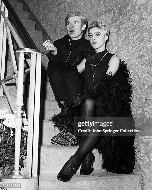 Artist Andy Warhol and Edie Sedgwick, who wears a black feather boa, sit on a staircase chatting. Undated photo.