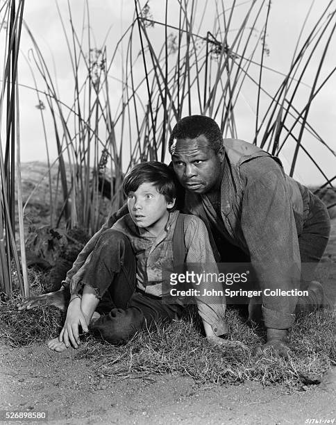 Huckleberry Finn and Jim crouch down along marsh weeds in a publicity still for The Adventures of Huckleberry Finn.