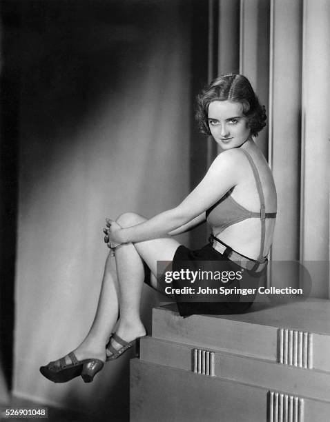 Portrait of Bette Davis sitting with her hands on her knees wearing a pair of strapped clogs, dark shorts, and a sexy sleeveless top. Undated...