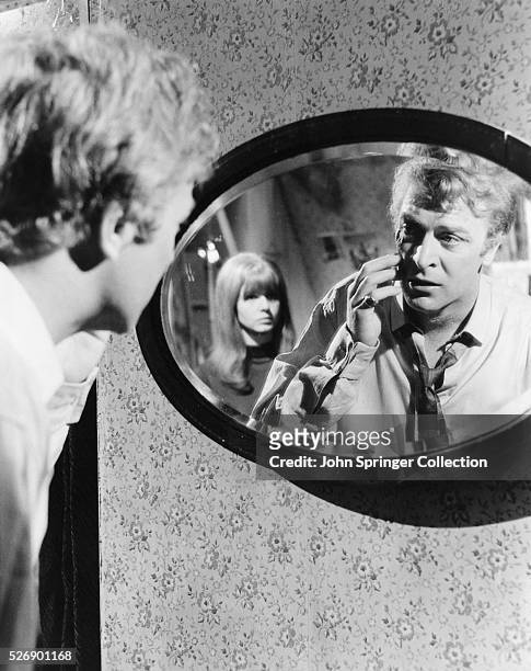 Michael Caine, portraying Alfie Elkins with Jane Asher in the 1966 motion picture Alfie, looks into a mirror.